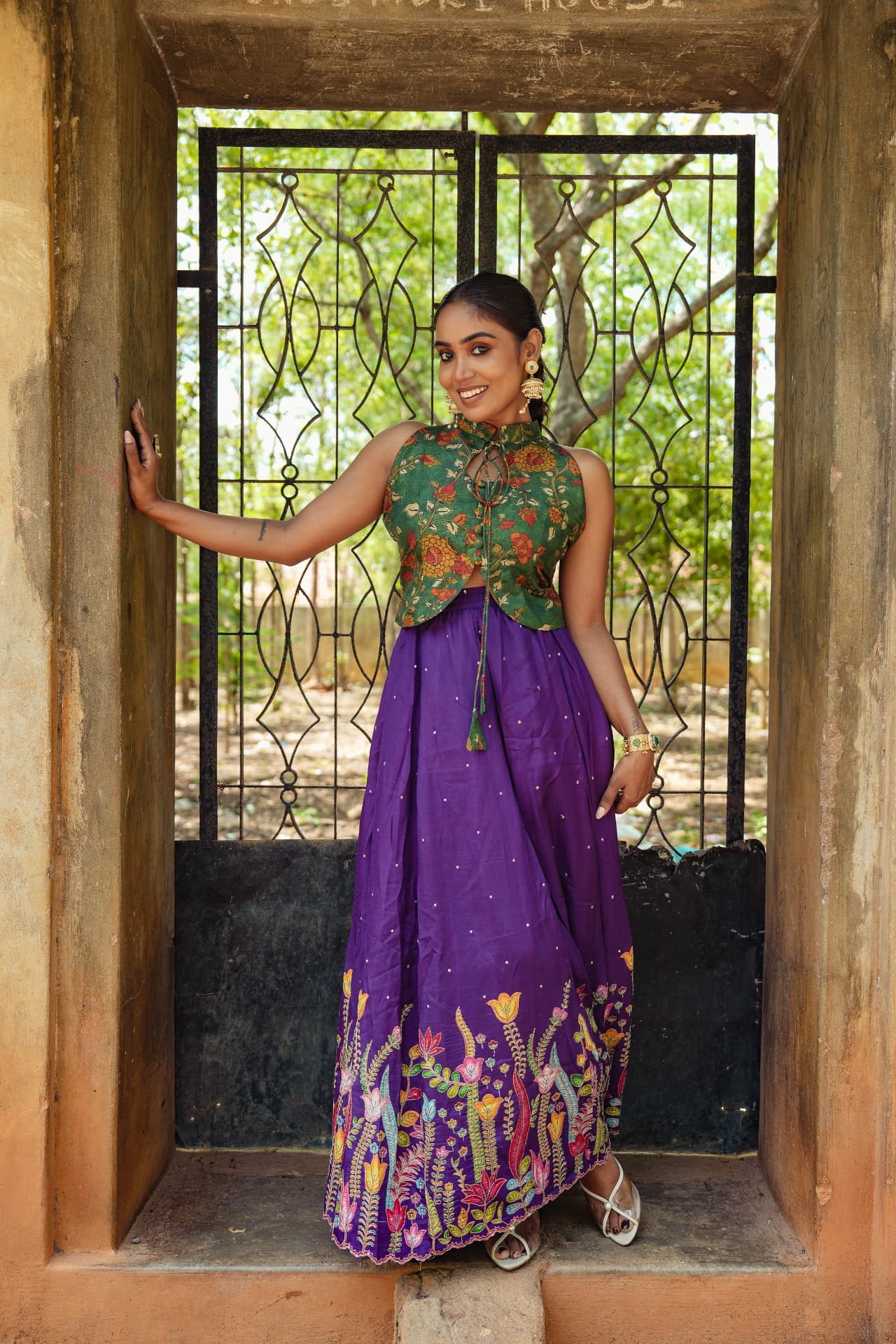 Model wearing a purple hand-embroidered floral skirt with a green kalamkari halter top in a heritage doorway.