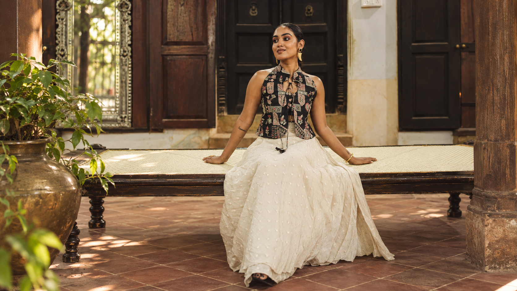 Model in an ivory pearl-work lehenga with a printed halter, seated on a carved daybed in a tiled courtyard.